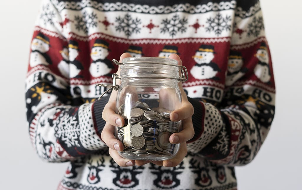 A man in a winter-themed sweater holds a school fundraising jar full of coins.