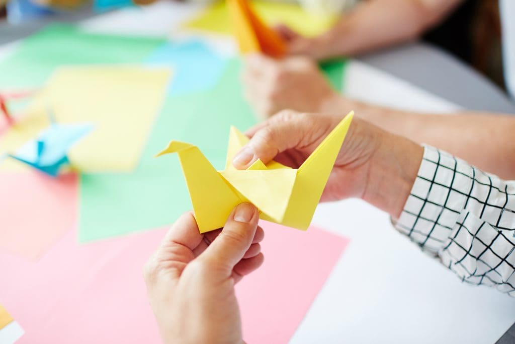 A teacher sits at a school table folding a yellow paper dove for a fundraising drive.