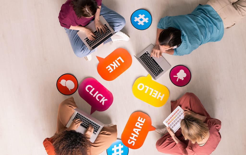 Students sitting on the floor looking at laptops surrounded by cutouts of words and icons representing social media engagement